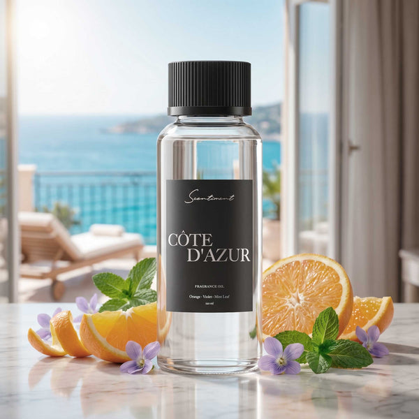 Scentiment Côte d’Azur fragrance oil bottle on a marble table, surrounded by orange slices, mint leaves, and violet flowers, with an open balcony overlooking a bright blue Mediterranean-style coastline in the background.